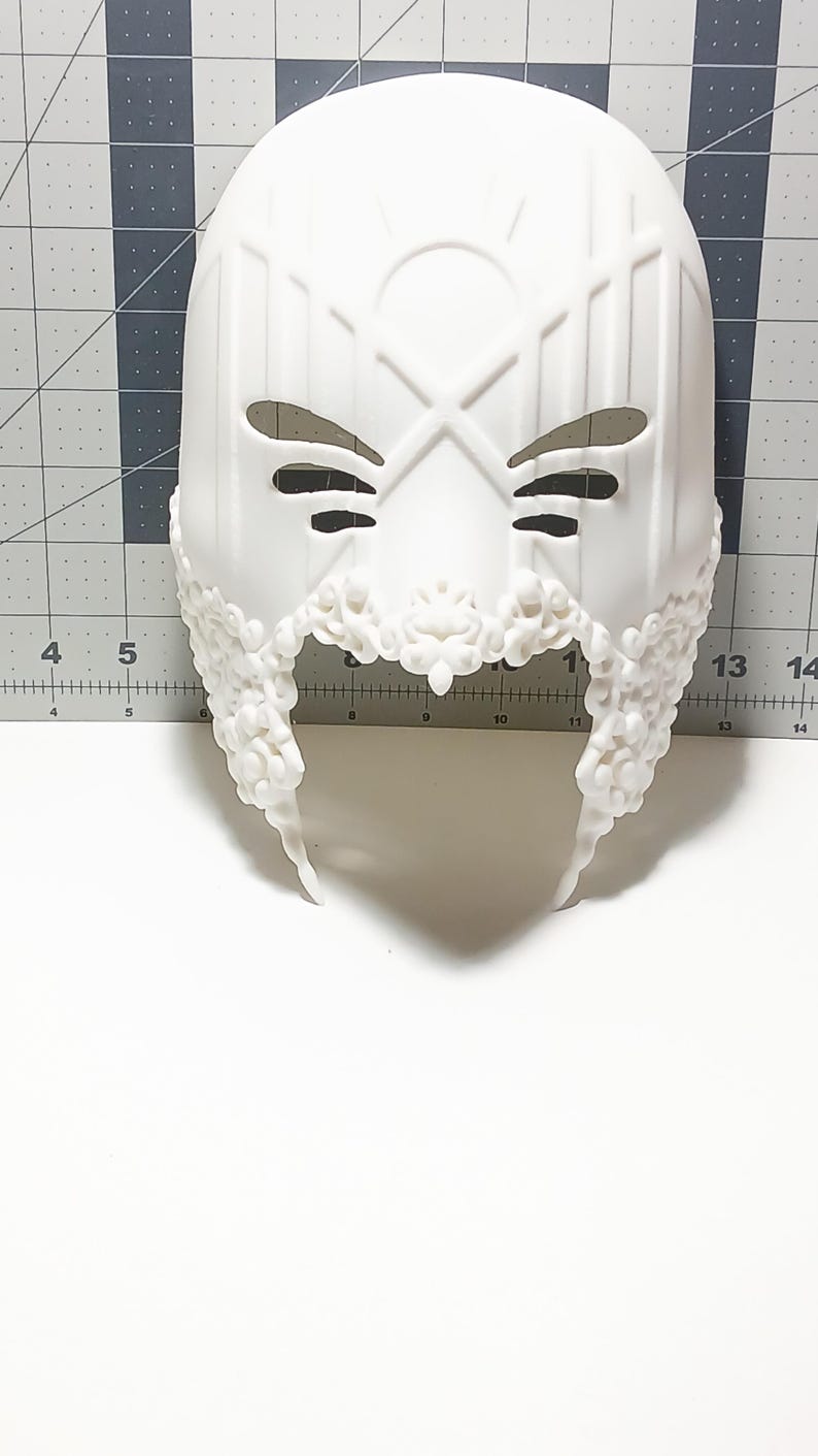 May include: White plastic mask with a detailed design. The mask has two eye holes and a decorative border around the bottom.