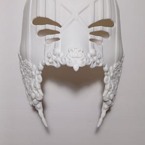May include: A white plastic mask with intricate details and a geometric design. The mask has two eye holes and a large, open mouth.