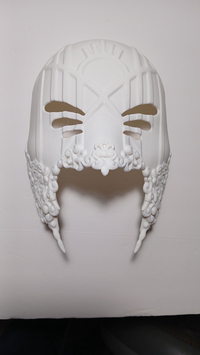 May include: A white plastic mask with intricate details and a geometric design. The mask has two eye holes and a large, open mouth.