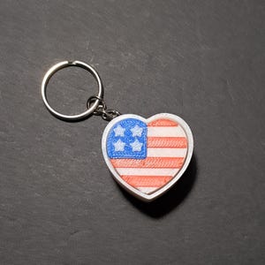 May include: A heart-shaped keychain featuring the American flag design. The keychain has a white border, a blue square with white stars, and red and white stripes. The keychain is attached to a silver keyring.