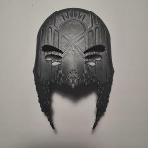 May include: A black 3D printed mask with intricate details and a geometric design. The mask has two eye holes and a pointed chin.