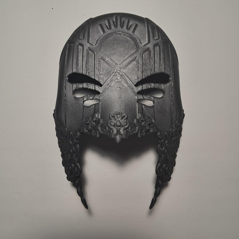 May include: A black 3D printed mask with intricate details and a geometric design. The mask has two eye holes and a pointed chin.