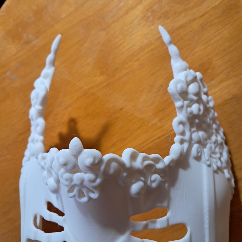 May include: White plastic masquerade mask with intricate floral details and a delicate, openwork design. The mask has a pointed top and a wide, open bottom.