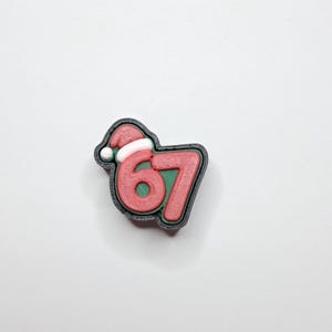 May include: A Christmas-themed charm featuring the number 67 in red with a green outline, a black border, and a Santa hat. The hat is red with a white brim and a white pom-pom. The charm is on a white background.