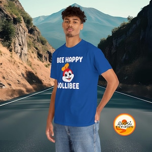 May include: A royal blue short-sleeved t-shirt featuring the white text BEE HAPPY and JOLLIBEE with a graphic of the Jollibee mascot wearing a chef hat. The shirt is worn with blue denim jeans, showcasing a casual style with a graphic print design.