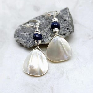 May include: A pair of dangle earrings featuring pearlescent, teardrop-shaped shells. Each earring has a dark blue bead and silver-toned metal accents. The earrings are displayed on a gray stone against a white background.