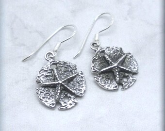 May include: Sterling silver earrings featuring a detailed sand dollar design with a starfish in the center. The earrings have a simple hook closure.