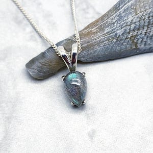 May include: A silver chain necklace with a teardrop-shaped pendant featuring a dark gray labradorite gemstone with iridescent blue and green flashes.