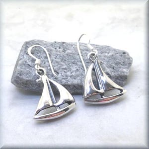 May include: A pair of silver sailboat earrings. Each earring features a detailed sailboat design with a curved hull, mast, and sail. The earrings have a hook closure and are displayed on a gray stone.