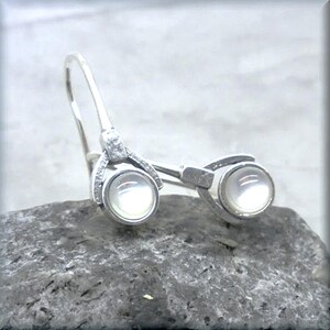 May include: A pair of silver earrings with white pearl-like stones. The earrings have a unique design with a curved shape and a small loop at the top.