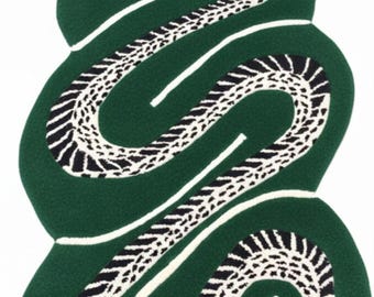 Puede incluir: Una alfombra de pelo insertado con forma de serpiente enroscada en color verde oscuro con un patrón blanco y negro en la parte inferior. La serpiente presenta un pequeño detalle naranja en la cabeza. El diseño es alargado y sigue la forma sinuosa.