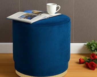 Puede incluir: Un otomán de terciopelo azul marino con base dorada. Un libro abierto y una taza blanca encima. El otomán es cilíndrico y tiene un aspecto suave y afelpado. Rosas rojas y vegetación en el fondo.