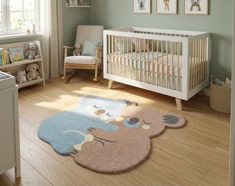 May include: A plush area rug shaped like two hugging koalas, one in light blue and one in light brown, featuring embroidered closed eyes and dark grey ears. The rug has a soft, textured pile and is positioned on a light-colored wooden floor.