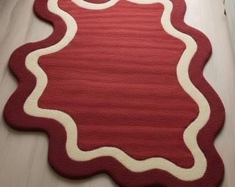 May include: An area rug with an irregular, organic shape featuring a deep red center and a thick, wavy cream-colored border. The rug has a textured, tufted surface and follows a fluid, non-symmetrical outline that curves along the edges.