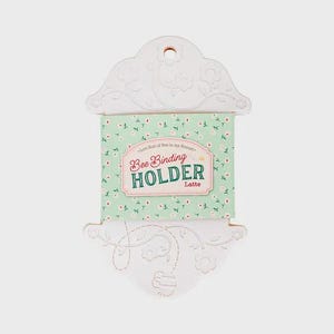 Peut inclure: Un porte-fil à coudre blanc découpé avec des motifs floraux et d'abeilles. Le support comporte un insert à motifs floraux verts et blancs avec le texte "Bee Binding Holder Latte". Le support a un trou en haut pour l'accrocher.