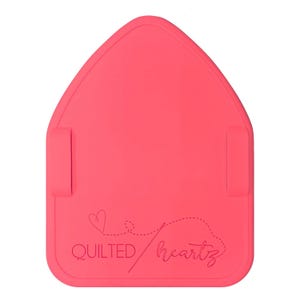 Op de afbeelding: Een felroze, huisvormige naaimat met twee zijlipjes. De mat heeft de tekst "QUILTED/heartz" met een hartje en een stippellijnontwerp. De mat is ontworpen voor quilt- en naaiprojecten.