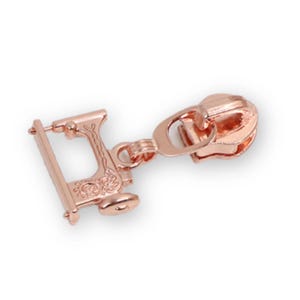 May include: A rose gold metal zipper pull featuring a detailed charm shaped like a vintage sewing machine. The charm is attached to a standard zipper slider mechanism, showcasing a metallic finish with embossed decorative patterns on the frame.