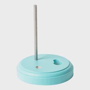 Puede incluir: Una base circular de madera de color azul claro con una varilla metálica plateada vertical que se extiende hacia arriba. La base presenta un acabado mate liso, una pequeña hendidura circular y un hueco en forma de cerradura.