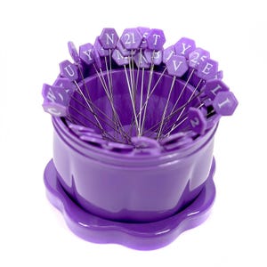 May include: A purple pin cushion with a scalloped base and a cylindrical body. The cushion is filled with numerous pins, each topped with a hexagonal purple head bearing a white letter or number. The pins are arranged around the edge of the cushion.
