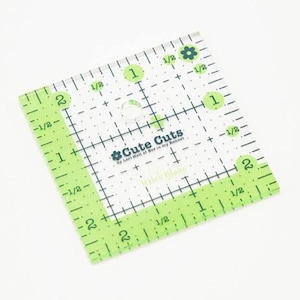 Op de afbeelding: Een doorzichtige, vierkante quiltliniaal met groene markeringen en de tekst "Cute Cuts". De liniaal heeft afmetingen in centimeters, inclusief 1/2 en 1 cm-stappen, en een bloemmotief.