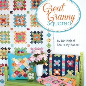 May include: A quilt pattern book titled Great Granny Squared by Lori Holt of Bee in my Bonnet, featuring a white quilt with colorful square blocks, a turquoise pillow, a navy quilt, a pink quilt, a green clock, and a white vase with flowers.