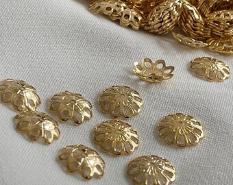 May include: Close-up of several gold-colored filigree bead caps, scattered on a white surface. The bead caps have a floral design with intricate cut-out patterns. Some caps are in focus, while others are slightly blurred.