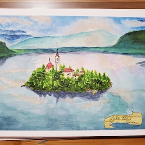 Può includere: Dipinto ad acquerello del lago di Bled in Slovenia. Il dipinto raffigura una piccola isola con una chiesa al centro del lago. Il lago è circondato da montagne e alberi. Il cielo è blu con nuvole bianche.