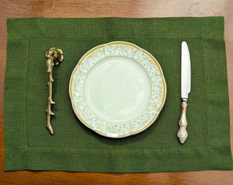 Puede incluir: Un mantel individual verde con un plato blanco con diseño floral, un cuchillo plateado y un utensilio de metal dorado en forma de rama.