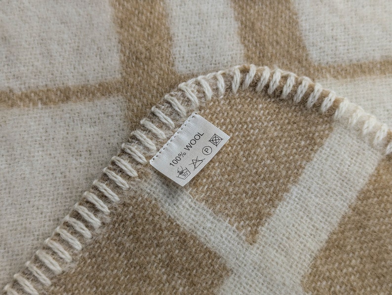 May include: A close-up of a beige and white checkered throw blanket with a white label that reads "100% Wool" and has care instructions.