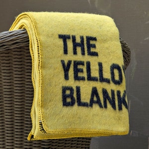 Puede incluir: Una manta amarilla de lana con texto negro que dice "THE YELLOW BLANKET". La manta está doblada y colocada sobre el respaldo de una silla de mimbre.