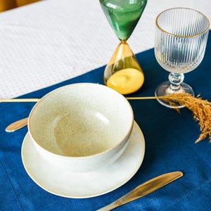 Puede incluir: Un cuenco de cer&aacute;mica blanca con un acabado moteado se asienta sobre un platillo blanco. El cuenco y el platillo est&aacute;n sobre un mantel individual azul. Un vaso de agua y un reloj de arena est&aacute;n sobre un mantel blanco con un patr&oacute;n de lunares.
