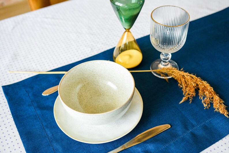 Puede incluir: Un cuenco de cer&aacute;mica blanca con un acabado moteado se asienta sobre un platillo blanco. El cuenco y el platillo est&aacute;n sobre un mantel individual azul. Un vaso de agua y un reloj de arena est&aacute;n sobre un mantel blanco con un patr&oacute;n de lunares.