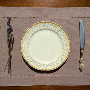 May include: A white ceramic dinner plate with a floral pattern in shades of yellow, orange, and blue. The plate is set on a brown linen placemat with a single sprig of lavender.