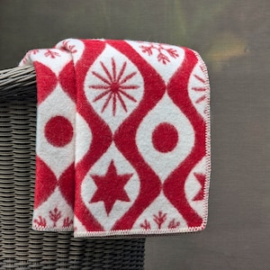 Puede incluir: Una manta plegada con un patr&oacute;n rojo y blanco, con dise&ntilde;os de estrellas, c&iacute;rculos y formas de copo de nieve. La manta est&aacute; sobre una silla de mimbre, mostrando su intrincado patr&oacute;n y combinaci&oacute;n de colores. Medidas no visibles.
