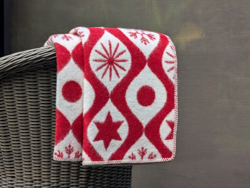Puede incluir: Una manta plegada con un patr&oacute;n rojo y blanco, con dise&ntilde;os de estrellas, c&iacute;rculos y formas de copo de nieve. La manta est&aacute; sobre una silla de mimbre, mostrando su intrincado patr&oacute;n y combinaci&oacute;n de colores. Medidas no visibles.