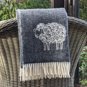 May include: A dark gray wool throw blanket with a white sheep design. The blanket has a fringe border.