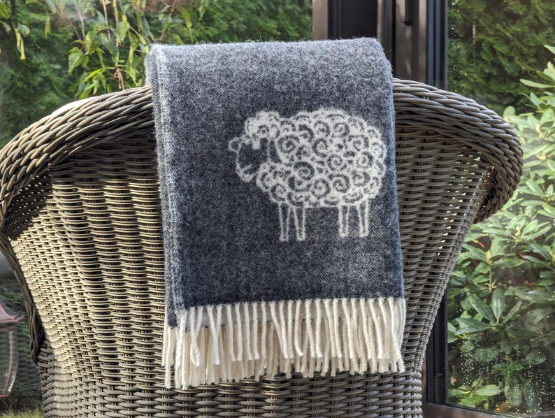 May include: A dark gray wool throw blanket with a white sheep design. The blanket has a fringe border.