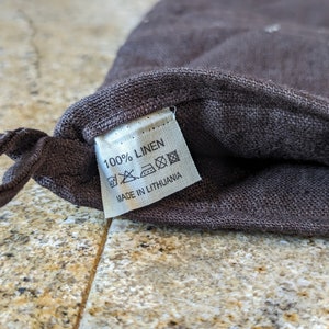 K&ouml;nnte beinhalten: Eine braune Leinentasche mit einem wei&szlig;en Etikett, auf dem "100% LINEN" und "MADE IN LITHUANIA" steht. Das Etikett enth&auml;lt Pflegehinweise mit Symbolen f&uuml;r Maschinenw&auml;sche, Bleiche, Trocknergeeignet und B&uuml;geln.