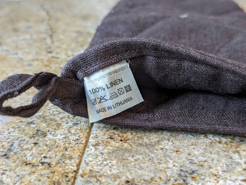 K&ouml;nnte beinhalten: Eine braune Leinentasche mit einem wei&szlig;en Etikett, auf dem "100% LINEN" und "MADE IN LITHUANIA" steht. Das Etikett enth&auml;lt Pflegehinweise mit Symbolen f&uuml;r Maschinenw&auml;sche, Bleiche, Trocknergeeignet und B&uuml;geln.