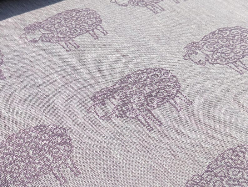 May include: A white fabric with a repeating pattern of purple sheep. The sheep are stylised and have a curly wool design.