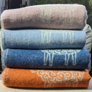 May include: Four folded blankets stacked on a wicker chair. The blankets are in shades of pink, blue, denim blue, and orange. Each blanket has a white sheep design.