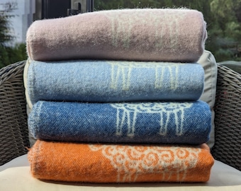 May include: Four folded blankets stacked on a wicker chair. The blankets are in shades of pink, blue, denim blue, and orange. Each blanket has a white sheep design.