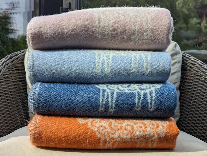 May include: Four folded blankets stacked on a wicker chair. The blankets are in shades of pink, blue, denim blue, and orange. Each blanket has a white sheep design.