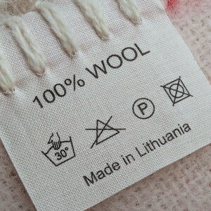 May include: A white fabric label with black text that reads "100% WOOL" and "Made in Lithuania". The label also includes several care instructions symbols, including a hand wash symbol with a 30 degree Celsius temperature, a do not iron symbol, a do not dry clean symbol, and a do not bleach symbol.