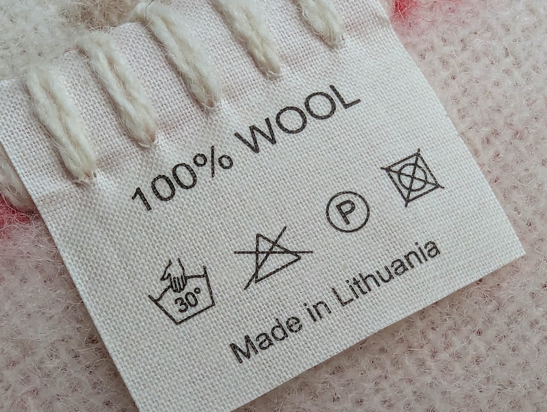 May include: A white fabric label with black text that reads "100% WOOL" and "Made in Lithuania". The label also includes several care instructions symbols, including a hand wash symbol with a 30 degree Celsius temperature, a do not iron symbol, a do not dry clean symbol, and a do not bleach symbol.