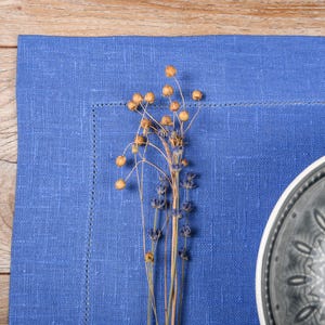 May include: A blue linen placemat with a gray ceramic plate with a decorative design. A sprig of dried flowers with brown seed pods is in the center of the placemat.