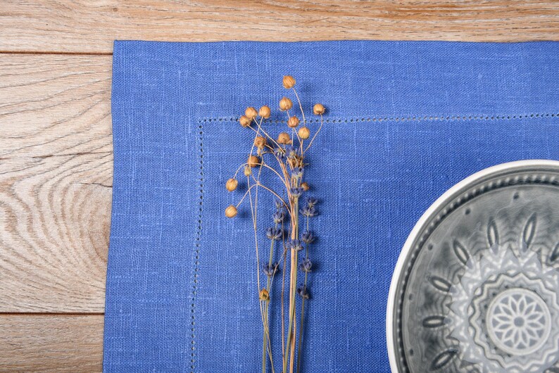 May include: A blue linen placemat with a gray ceramic plate with a decorative design. A sprig of dried flowers with brown seed pods is in the center of the placemat.