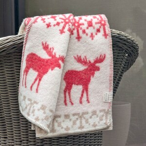 May include: A white and red throw blanket with a Scandinavian-style pattern featuring moose. The blanket is folded and draped over the back of a wicker chair.