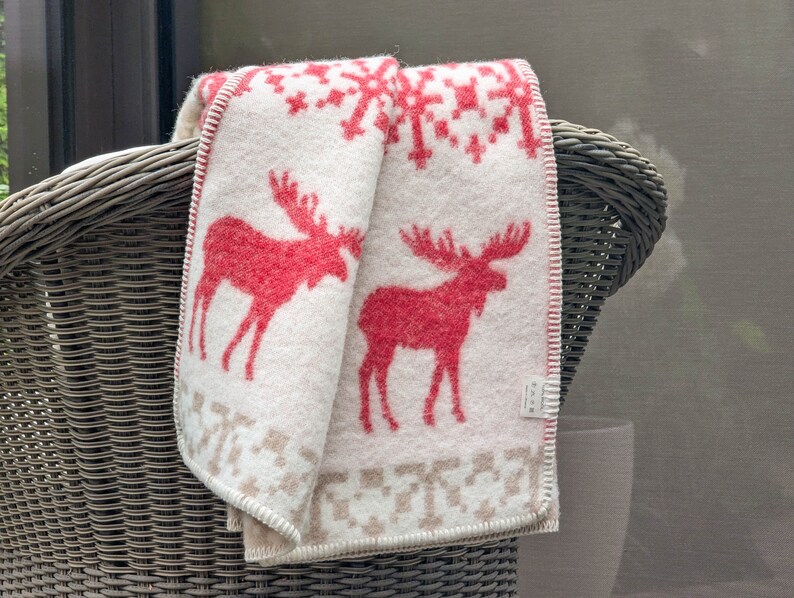May include: A white and red throw blanket with a Scandinavian-style pattern featuring moose. The blanket is folded and draped over the back of a wicker chair.