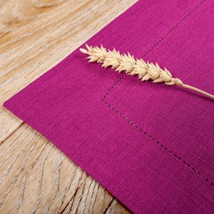 May include: A close-up of a purple linen placemat with a single stalk of wheat resting on top.